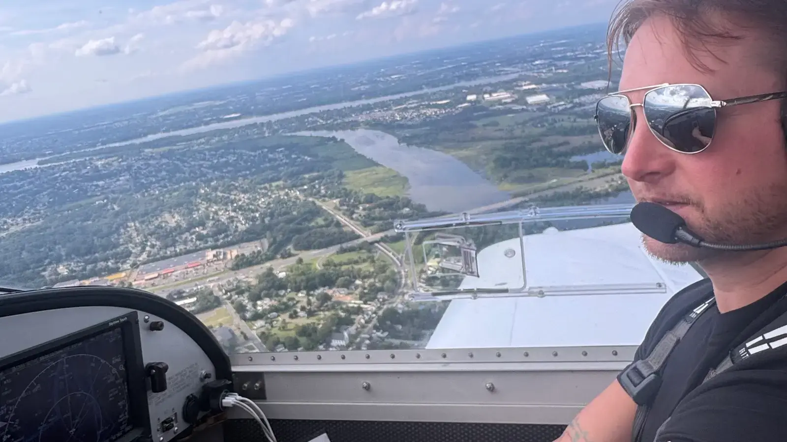A pilot in a headset and sunglasses at the controls of N159G, with towns and a river below